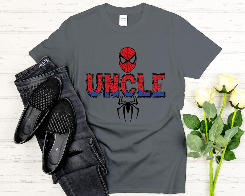 Może przedstawiać: Szara koszulka z motywem Spider-Mana. Słowo "UNCLE" jest wydrukowane w kolorze czerwonym i niebieskim, z maską Spider-Mana powyżej i grafiką pająka poniżej. Koszulka jest połączona z czarnymi dżinsami i butami.