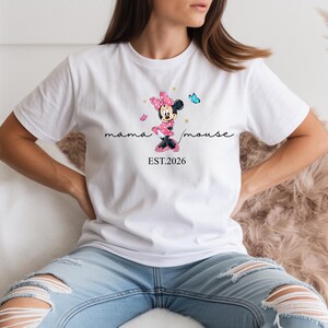 May include: White t-shirt with a Minnie Mouse graphic in pink and black, the text reads "mama mouse" and "EST.2026". The design includes pink butterflies. A casual wear item.