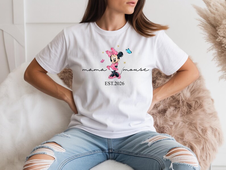 May include: White t-shirt with a Minnie Mouse graphic in pink and black, the text reads "mama mouse" and "EST.2026". The design includes pink butterflies. A casual wear item.