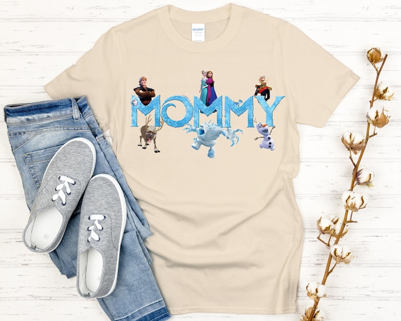 May include: Beige t-shirt with the word "MOMMY" in blue, featuring characters from the movie Frozen. The shirt includes images of Anna, Elsa, Kristoff, Sven, Olaf, and Marshmallow. The shirt is paired with blue jeans and gray sneakers.