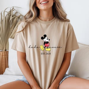 May include: A beige t-shirt with a Mickey Mouse graphic. The design includes the text "dada mouse" and "EST.2024". Mickey is depicted in his classic outfit: red shorts, yellow shoes, and black gloves.