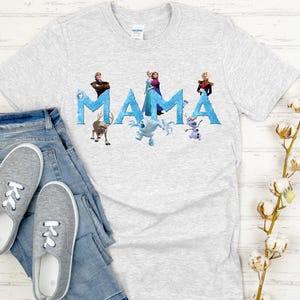 May include: A light gray t-shirt with the word "MAMA" in blue letters, featuring characters from the movie Frozen. The shirt is paired with blue jeans and gray sneakers. The shirt is made of a soft material.