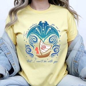 Op de afbeelding: Lichtgeel T-shirt met een grafisch ontwerp met een boot met een wit zeil, een silhouet van een persoon en wervelende blauwe golven. Het ontwerp bevat de tekst "There is nowhere you can go that I won't be with you..."