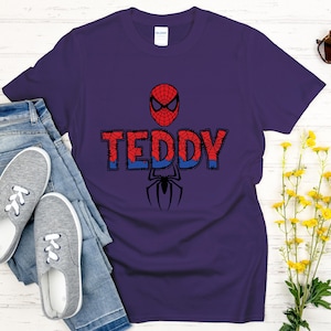 Może przedstawiać: Fioletowa koszulka z motywem Spider-Mana. Projekt zawiera maskę postaci, imię "TEDDY" w kolorze czerwonym i niebieskim oraz czarną sylwetkę pająka. Koszulka jest zestawiona z niebieskimi dżinsami i szarymi trampkami.