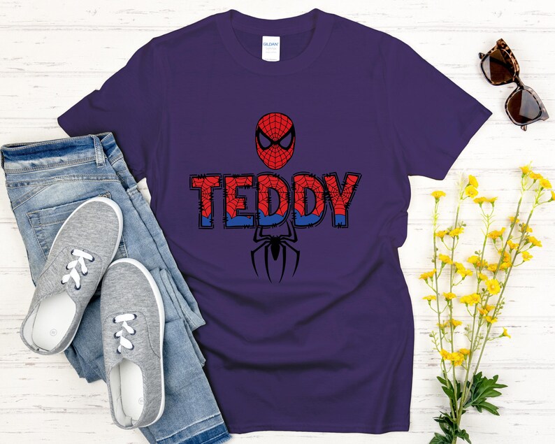 Może przedstawiać: Fioletowa koszulka z motywem Spider-Mana. Projekt zawiera maskę postaci, imię "TEDDY" w kolorze czerwonym i niebieskim oraz czarną sylwetkę pająka. Koszulka jest zestawiona z niebieskimi dżinsami i szarymi trampkami.