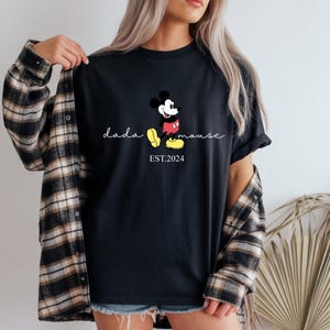 May include: Black t-shirt with a Mickey Mouse graphic and the text "dada mouse EST. 2024". The design shows Mickey in his classic pose with red shorts, yellow shoes, and white gloves. The shirt is worn with a plaid flannel shirt.