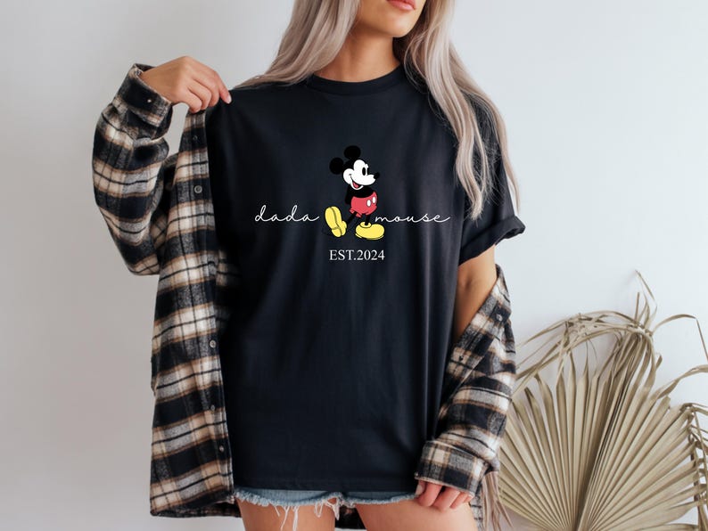 May include: Black t-shirt with a Mickey Mouse graphic and the text "dada mouse EST. 2024". The design shows Mickey in his classic pose with red shorts, yellow shoes, and white gloves. The shirt is worn with a plaid flannel shirt.