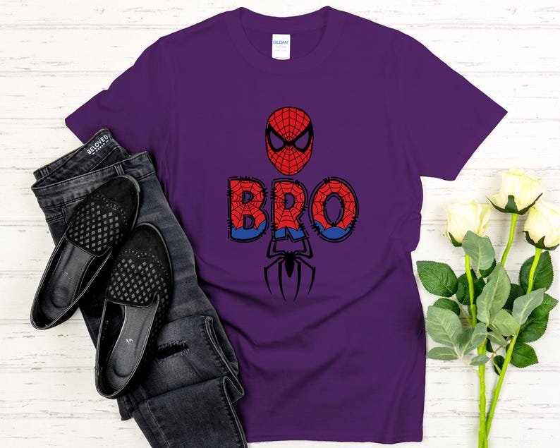 Może przedstawiać: Fioletowa koszulka z motywem Spider-Mana. Słowo "BRO" jest w kolorze czerwonym i niebieskim, z maską Spider-Mana powyżej i grafiką pająka poniżej. Koszulka jest zestawiona z czarnymi dżinsami i butami.
