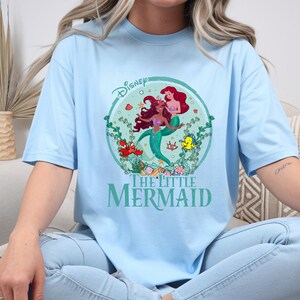K&ouml;nnte beinhalten: Hellblaues T-Shirt mit einem kreisf&ouml;rmigen Aufdruck von Arielle und anderen Figuren aus Disneys &bdquo;Arielle, die Meerjungfrau&ldquo;. Die Grafik enth&auml;lt das Disney-Logo und die Worte &bdquo;The Little Mermaid&ldquo; in Gr&uuml;n. Das Shirt hat eine lockere Passform.