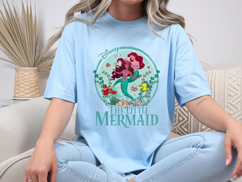 K&ouml;nnte beinhalten: Hellblaues T-Shirt mit einem kreisf&ouml;rmigen Aufdruck von Arielle und anderen Figuren aus Disneys &bdquo;Arielle, die Meerjungfrau&ldquo;. Die Grafik enth&auml;lt das Disney-Logo und die Worte &bdquo;The Little Mermaid&ldquo; in Gr&uuml;n. Das Shirt hat eine lockere Passform.