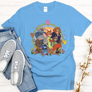 Peut inclure: Un t-shirt bleu clair avec un graphisme color&eacute; de personnages de dessins anim&eacute;s. Le motif comprend des personnages d'animaux, des fleurs et des ballons. Le t-shirt est pr&eacute;sent&eacute; avec un jean bleu et des baskets grises.
