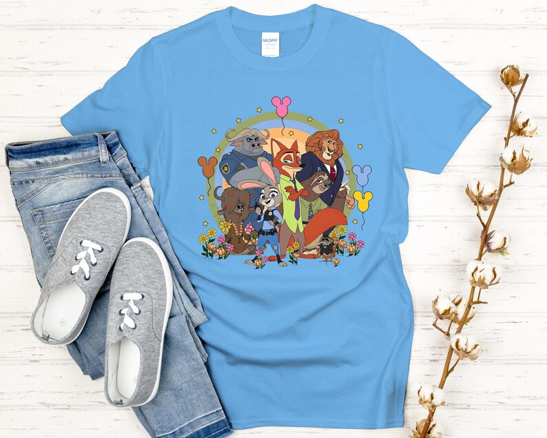 Peut inclure: Un t-shirt bleu clair avec un graphisme color&eacute; de personnages de dessins anim&eacute;s. Le motif comprend des personnages d'animaux, des fleurs et des ballons. Le t-shirt est pr&eacute;sent&eacute; avec un jean bleu et des baskets grises.