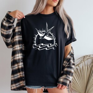 K&ouml;nnte beinhalten: Schwarzes T-Shirt mit wei&szlig;er Grafik einer Schwalbe vor Sonne und Wellen. Das Shirt wird mit einem karierten Flanellhemd getragen. Das Design ist im nautischen oder Vintage-Stil gehalten.