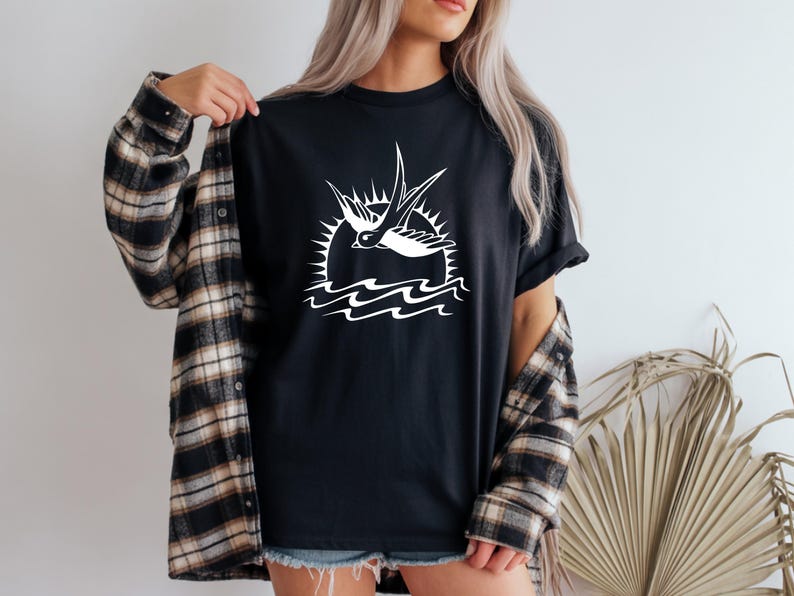 K&ouml;nnte beinhalten: Schwarzes T-Shirt mit wei&szlig;er Grafik einer Schwalbe vor Sonne und Wellen. Das Shirt wird mit einem karierten Flanellhemd getragen. Das Design ist im nautischen oder Vintage-Stil gehalten.
