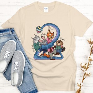 Puede incluir: Una camiseta beige con un dibujo animado colorido de personajes de animales, incluyendo un zorro, un conejo y un castor, con el n&uacute;mero "2" y nombres de personajes. La camiseta se muestra con vaqueros azules y zapatillas grises.