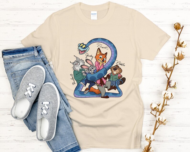 Puede incluir: Una camiseta beige con un dibujo animado colorido de personajes de animales, incluyendo un zorro, un conejo y un castor, con el n&uacute;mero "2" y nombres de personajes. La camiseta se muestra con vaqueros azules y zapatillas grises.
