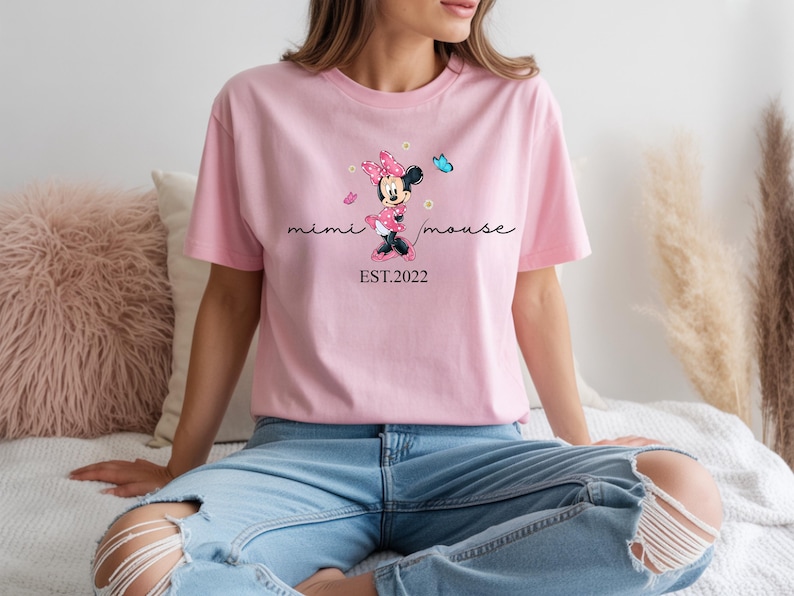 May include: A pale pink t-shirt with a Minnie Mouse design, including a pink bow and a blue butterfly. The words "mimi mouse" are in cursive, with "EST.2022" underneath. The shirt is worn with blue jeans.