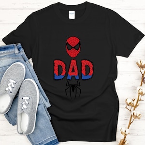 Może przedstawiać: Czarny t-shirt z czerwono-niebieskim wzorem Spider-Mana i napisem "DAD". Projekt zawiera maskę Spider-Mana, słowo "DAD" w stylu komiksowym i sylwetkę pająka. Koszulka jest połączona z niebieskimi dżinsami i szarymi trampkami.