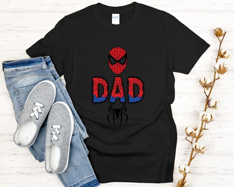 Może przedstawiać: Czarny t-shirt z czerwono-niebieskim wzorem Spider-Mana i napisem "DAD". Projekt zawiera maskę Spider-Mana, słowo "DAD" w stylu komiksowym i sylwetkę pająka. Koszulka jest połączona z niebieskimi dżinsami i szarymi trampkami.