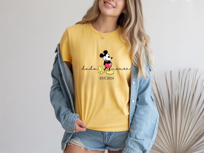 May include: Mustard yellow t-shirt with a Mickey Mouse design, the words "dada mouse" and "EST.2024". The shirt is worn with denim shorts and a light blue denim jacket.