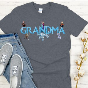 May include: A gray t-shirt with the word "GRANDMA" in blue, featuring cartoon characters. The shirt is paired with blue jeans and gray sneakers. A sprig of cotton flowers is in the background.