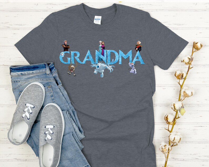 May include: A gray t-shirt with the word "GRANDMA" in blue, featuring cartoon characters. The shirt is paired with blue jeans and gray sneakers. A sprig of cotton flowers is in the background.
