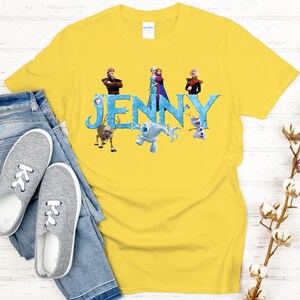 May include: A yellow t-shirt featuring the name "JENNY" in blue letters with characters from a popular animated movie. The shirt is paired with blue jeans and gray sneakers, creating a casual, everyday look.