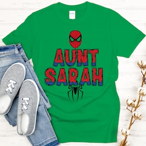 Może przedstawiać: Zielona koszulka z napisem "AUNT SARAH" w kolorze czerwonym i niebieskim, z maską Spider-Mana i grafiką pająka. Koszulka jest zestawiona z niebieskimi dżinsami i szarymi trampkami.