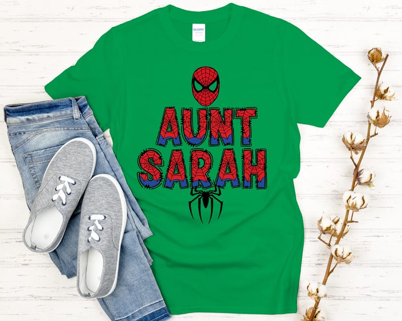 Może przedstawiać: Zielona koszulka z napisem "AUNT SARAH" w kolorze czerwonym i niebieskim, z maską Spider-Mana i grafiką pająka. Koszulka jest zestawiona z niebieskimi dżinsami i szarymi trampkami.
