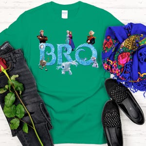 May include: A green t-shirt featuring the word "BRO" in icy blue letters, with characters from the movie Frozen. The shirt is paired with black jeans, a red rose, a blue scarf with floral patterns, and black shoes.