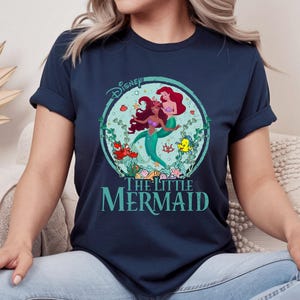 K&ouml;nnte beinhalten: Marineblaues T-Shirt mit einem farbenfrohen Grafikdesign von Disneys "Arielle, die Meerjungfrau". Das Design zeigt Arielle und eine weitere Meerjungfrau, umgeben von Wasserpflanzen und Meeresbewohnern. Der Text "The Little Mermaid" ist darunter gedruckt.