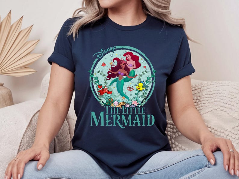 K&ouml;nnte beinhalten: Marineblaues T-Shirt mit einem farbenfrohen Grafikdesign von Disneys "Arielle, die Meerjungfrau". Das Design zeigt Arielle und eine weitere Meerjungfrau, umgeben von Wasserpflanzen und Meeresbewohnern. Der Text "The Little Mermaid" ist darunter gedruckt.