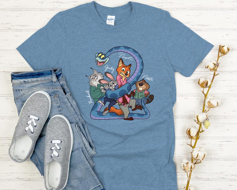 Puede incluir: Una camiseta azul claro con un dibujo animado de personajes de una pel&iacute;cula. El gr&aacute;fico incluye una serpiente y varios personajes de animales, con los nombres Judy, Nick y otros. La camiseta se combina con vaqueros azules y zapatillas grises.