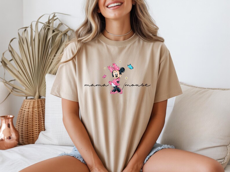 May include: A beige t-shirt with a Minnie Mouse graphic in a pink bow and dress, and the text "mama mouse". The design includes a blue butterfly and pink hearts. The shirt is a neutral color, suitable for various outfits.