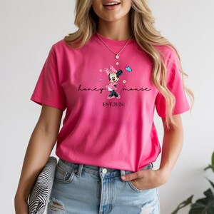 May include: A vibrant pink t-shirt with a Minnie Mouse graphic and the text "honey mouse EST. 2024". The design includes a cartoon Minnie Mouse, butterflies, and script lettering. The shirt is a short-sleeved crew neck.
