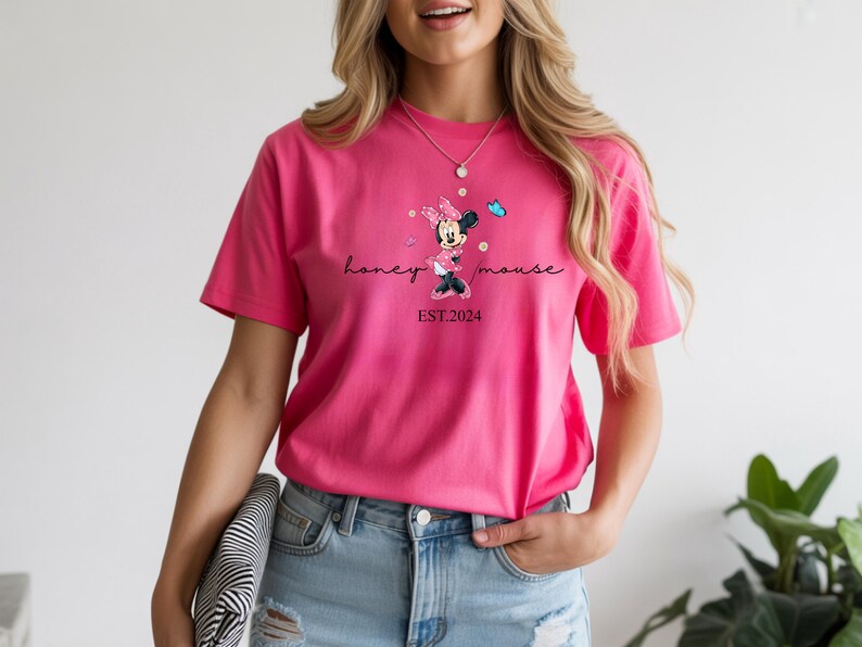 May include: A vibrant pink t-shirt with a Minnie Mouse graphic and the text "honey mouse EST. 2024". The design includes a cartoon Minnie Mouse, butterflies, and script lettering. The shirt is a short-sleeved crew neck.