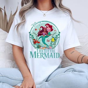 K&ouml;nnte beinhalten: Wei&szlig;es T-Shirt mit einer farbenfrohen Grafik von Arielle und anderen Charakteren aus Disneys Arielle, die Meerjungfrau. Das Design befindet sich in einem kreisf&ouml;rmigen Rahmen mit dem Disney-Logo oben. Die Worte "The Little Mermaid" sind unten aufgedruckt.
