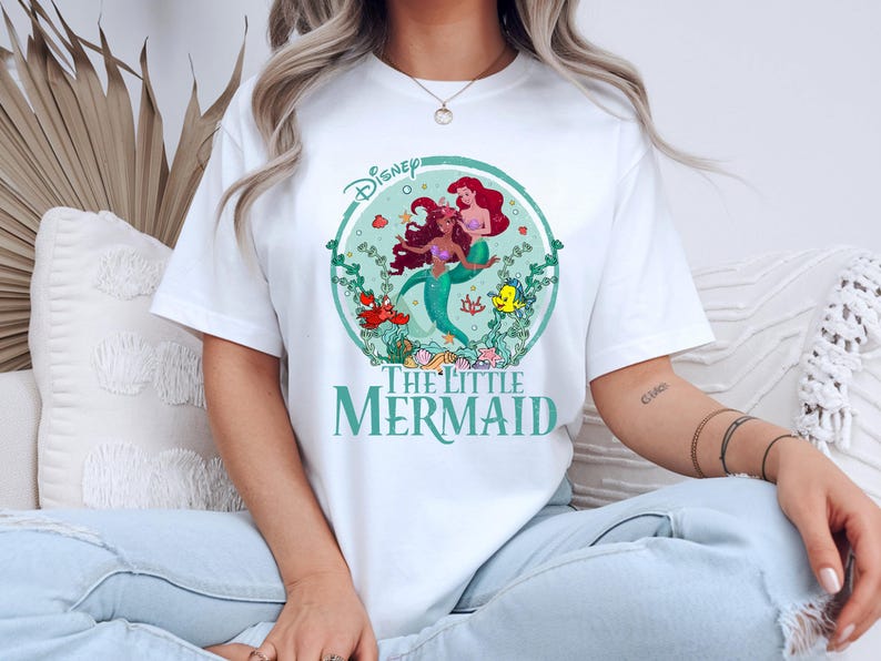 K&ouml;nnte beinhalten: Wei&szlig;es T-Shirt mit einer farbenfrohen Grafik von Arielle und anderen Charakteren aus Disneys Arielle, die Meerjungfrau. Das Design befindet sich in einem kreisf&ouml;rmigen Rahmen mit dem Disney-Logo oben. Die Worte "The Little Mermaid" sind unten aufgedruckt.
