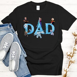 May include: Black t-shirt with the word "DAD" in blue, featuring characters from the movie Frozen. The shirt is paired with blue jeans and gray sneakers. A sprig of cotton adds a decorative touch.