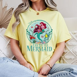 K&ouml;nnte beinhalten: Hellgelbes T-Shirt mit einem farbenfrohen Grafikdesign von Arielle und anderen Charakteren aus Disneys Arielle, die Meerjungfrau. Das Design ist in einem Kreis mit dem Filmtitel in T&uuml;rkis eingefasst. Das T-Shirt besteht aus weichem Material.