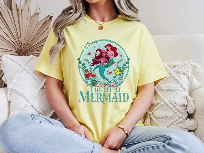 K&ouml;nnte beinhalten: Hellgelbes T-Shirt mit einem farbenfrohen Grafikdesign von Arielle und anderen Charakteren aus Disneys Arielle, die Meerjungfrau. Das Design ist in einem Kreis mit dem Filmtitel in T&uuml;rkis eingefasst. Das T-Shirt besteht aus weichem Material.
