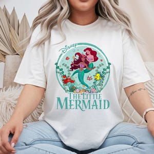 K&ouml;nnte beinhalten: Off-White-T-Shirt mit einem teal- und roten Grafik von Arielle und anderen Charakteren aus Disneys The Little Mermaid. Das Disney-Logo befindet sich oben, und die Worte "The Little Mermaid" stehen unten in Teal.