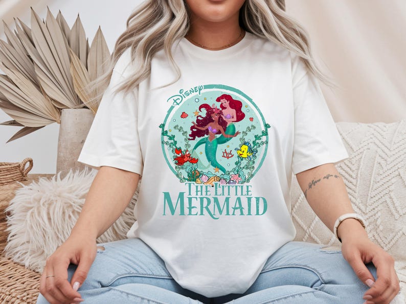 K&ouml;nnte beinhalten: Off-White-T-Shirt mit einem teal- und roten Grafik von Arielle und anderen Charakteren aus Disneys The Little Mermaid. Das Disney-Logo befindet sich oben, und die Worte "The Little Mermaid" stehen unten in Teal.