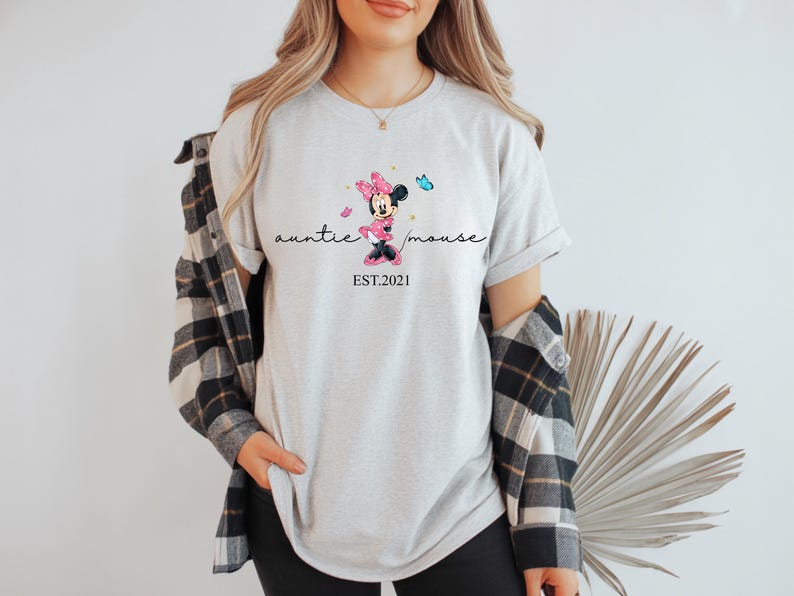 May include: A light grey t-shirt with a Minnie Mouse graphic, the text reads "auntie mouse" and "EST. 2021". The graphic shows Minnie Mouse in a pink dress and bow, with butterflies. The shirt is a casual style.