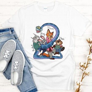 Puede incluir: Camiseta blanca con un gr&aacute;fico de dibujos animados colorido de personajes, incluyendo un zorro, un conejo y un castor, con los nombres Judy, Nick y Nibbles. El dise&ntilde;o est&aacute; sobre una forma azul en forma de serpiente. La camiseta se combina con vaqueros azules y zapatillas grises.