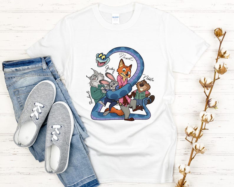 Puede incluir: Camiseta blanca con un gr&aacute;fico de dibujos animados colorido de personajes, incluyendo un zorro, un conejo y un castor, con los nombres Judy, Nick y Nibbles. El dise&ntilde;o est&aacute; sobre una forma azul en forma de serpiente. La camiseta se combina con vaqueros azules y zapatillas grises.