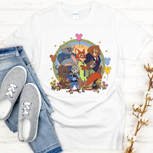 Peut inclure: T-shirt blanc avec une illustration de dessin anim&eacute; color&eacute;e de personnages d'un film d'animation populaire. Le motif comprend un lapin, un renard, un lion et d'autres personnages, entour&eacute;s de fleurs et de ballons. Le t-shirt est pr&eacute;sent&eacute; avec un jean bleu et des baskets grises.
