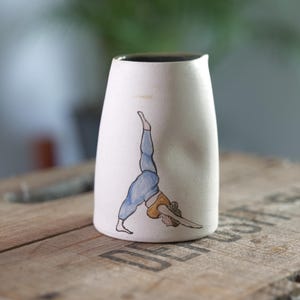 Könnte beinhalten: Weiße Keramikvase mit schwarzem Rand, mit einer handgemalten Illustration einer Person in einer Yoga-Pose. Die Figur trägt eine blaue Hose und ein gelbes Oberteil. Die Vase steht auf einer Holzoberfläche.