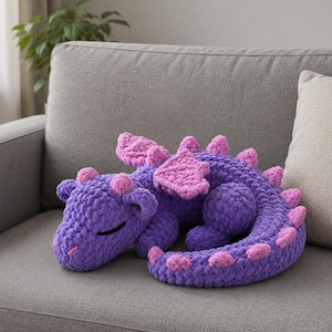 Puede incluir: Un dragón de peluche hecho de hilo grueso morado con suaves acentos rosas en las alas, espinas y hocico. La criatura está representada en una posición acurrucada y durmiente con los ojos cerrados, con una textura de tejido a ganchillo.