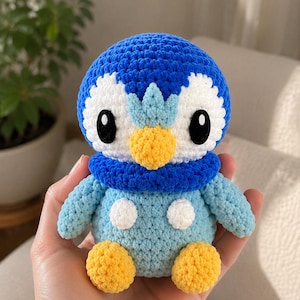 May include: A crochet plush toy shaped like a penguin with a blue head, white face patches, and a yellow beak. It features a dark blue neck collar, a light blue body with two white circular spots, and yellow feet. The eyes are large and black.
